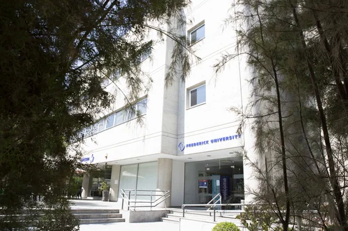 Entrance to a white multi-storey building with the university sign, partly framed by pine branches