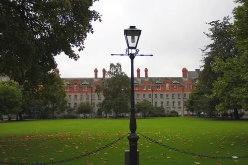 A lawn with fallen leaves, a lamp post and a chain fence in the foreground, and a stone and red-brick building with chimneys beyond the trees