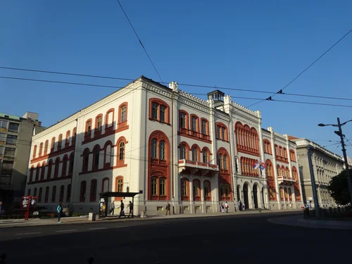 Pale building with red-framed arched windows on a street corner under trolleybus wires