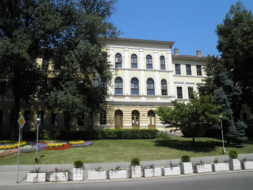 Pale building with arched windows behind trees and a flower bed by the road