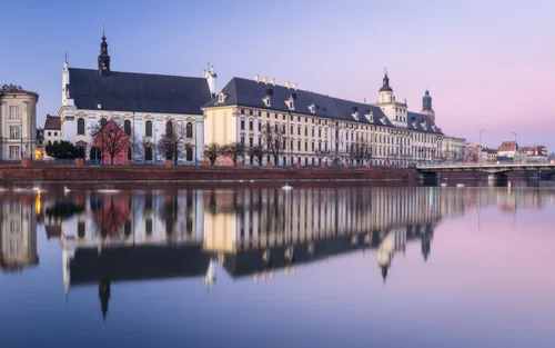 A long university facade and a church with a tall roof on the riverbank, the calm river reflecting the buildings at dusk