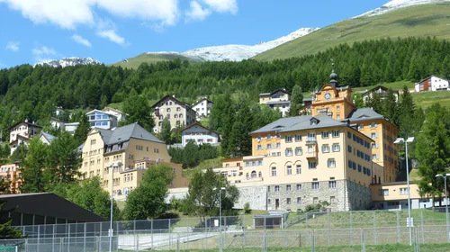 Large yellow buildings with a clock tower on a slope among spruce forest, with snow-capped mountains behind and a sports-ground fence in the foreground