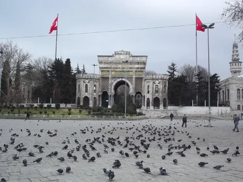 Moorish-style gate with clock turrets and an İstanbul Üniversitesi inscription, Turkish flags on either side and pigeons on the square