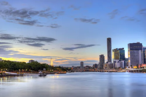 A calm river at dusk, with a lit riverside promenade and trees on the left and downtown towers and a bridge on the right