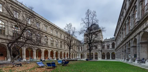 An inner courtyard with arcades around a lawn, blue deckchairs on fallen leaves, bare trees and a few people in the distance