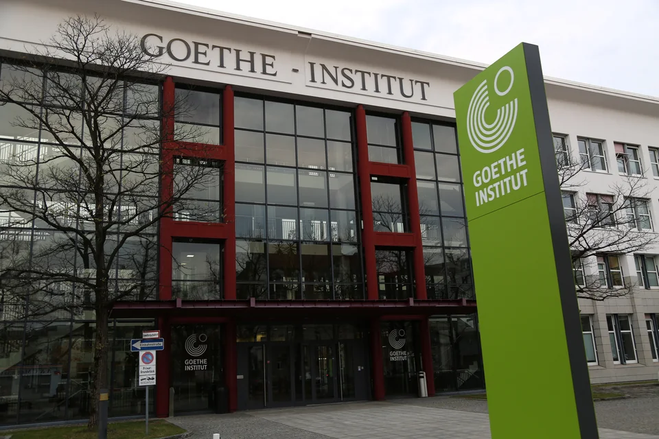 Светлое офисное здание со стеклянным фасадом в красных рамах и надписью Goethe-Institut, перед ним зелёная стела с логотипом