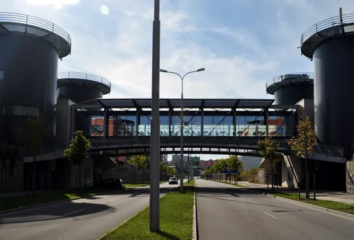 A glazed footbridge over a wide street between dark cylindrical buildings, with trees along the road