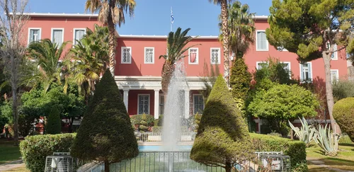 Red building with white columns, palms, a fountain and clipped conifers