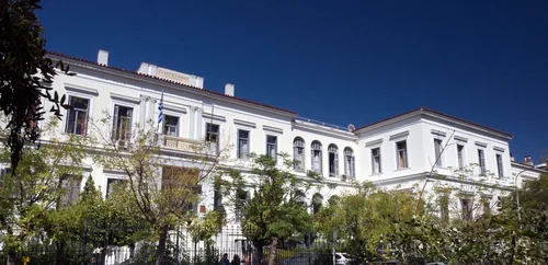 White neoclassical hospital building behind trees under a deep blue sky