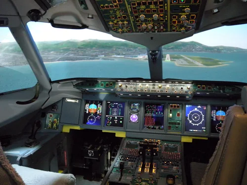 Airliner cockpit with digital displays and an overhead panel, a simulated approach to a seaside runway in the windows
