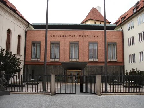 A brick façade with the words Universitas Carolina between pale side wings, with a metal fence and flagpoles in front