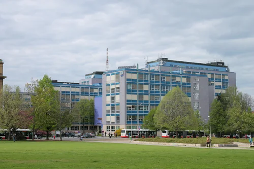 Multi-storey buildings with blue facades behind trees and a wide lawn under a spring sky