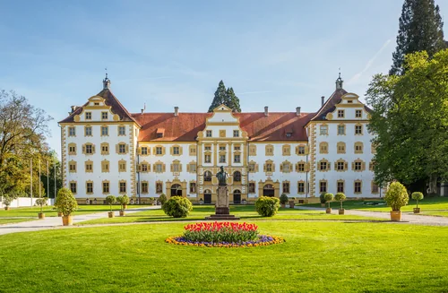A Baroque palace with a white and ochre façade and turrets, with a lawn, a statue and a tulip bed in front