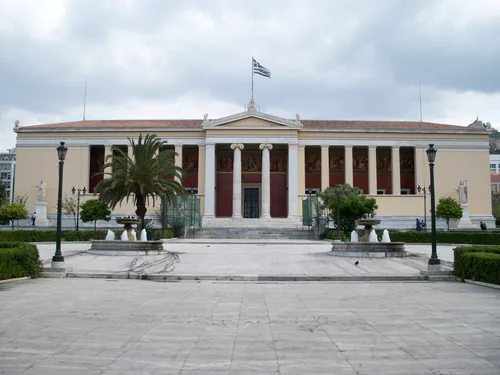 A neoclassical building with Ionic columns, a painted frieze and a Greek flag, with a palm tree, fountains and a marble square in front