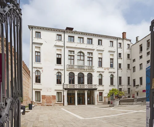A white palazzo with balconies and arched windows, an empty paved courtyard in front and a wrought-iron gate on the left