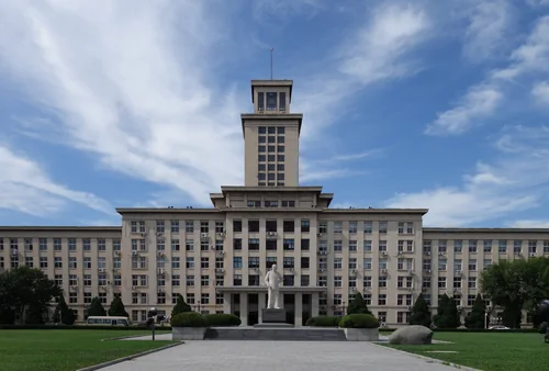 Tall main building with a central tower, a statue, lawn and trees in front