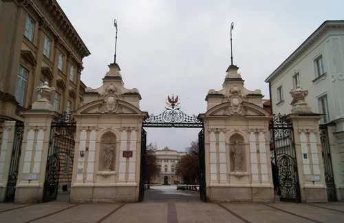 An ornate gateway with statues in niches and wrought ironwork topped by an eagle, with a palace at the end of the courtyard