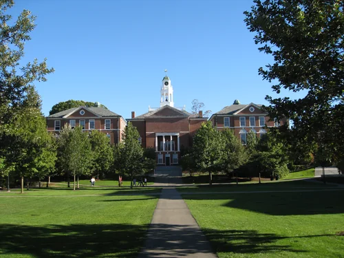 A brick building with a white cupola and two side wings, approached by a path across a lawn between trees