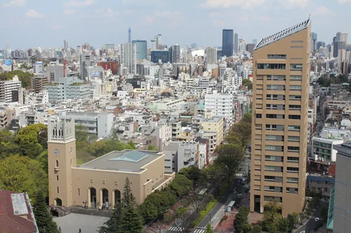 A view from above of a building with a clock tower and a tall beige block, surrounded by dense Tokyo city blocks