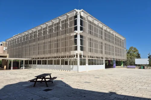Rectangular building with a mesh façade and an empty paved plaza with a picnic table