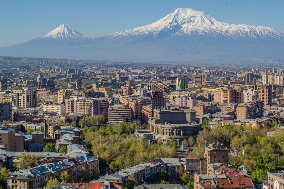 Панорама города с оперным театром среди зелени, за ней две заснеженные вершины