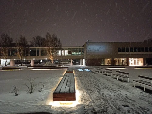 A low modernist building with lit windows and the sign Turun yliopisto, a snowy square with an illuminated bench