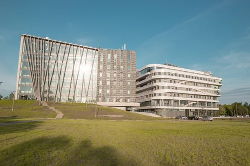 Two modern buildings, one glass with slanted supports and one white with rounded floors, beyond a lawn