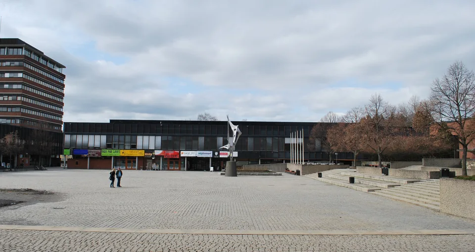 Широкая мощёная площадь перед длинным тёмным корпусом с деревьями по краям