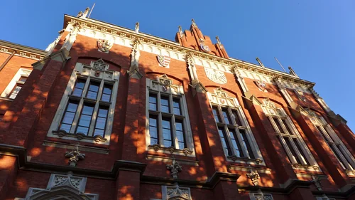 The facade of a red-brick Neo-Gothic building with tall windows and carved decoration, seen from below against a blue sky