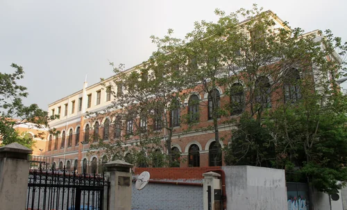 A red-brick building with arched windows behind tree canopies, with wrought-iron gates and a wall in the foreground