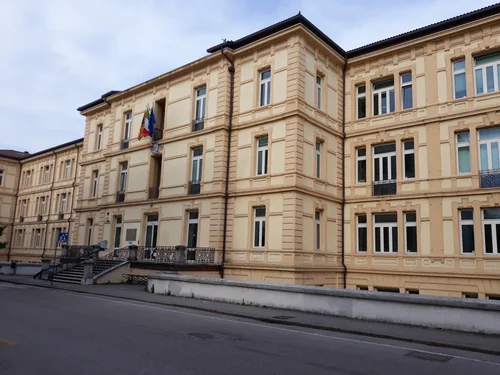 A three-storey school building with a beige façade, rows of windows and flags above the entrance, with a pavement and an empty street in front