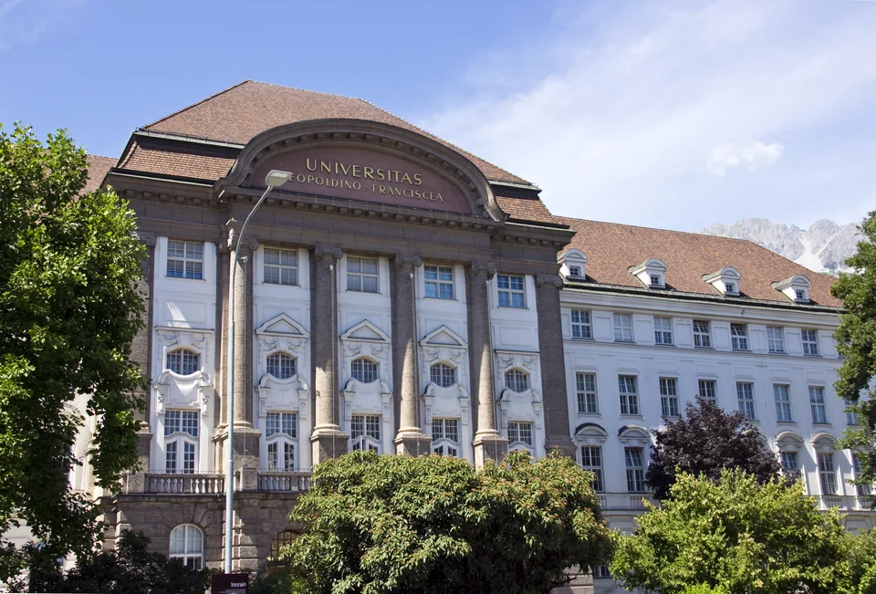 Фасад с надписью Universitas Leopoldino-Franciscea под черепичной крышей, вдали скалистые вершины