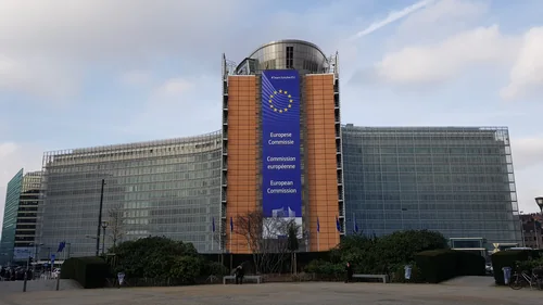 A large building with glass louvres and a European Commission banner with a ring of stars, with flags and shrubs in front