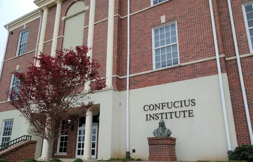 A brick building with white columns and the words Confucius Institute on the wall, with a bust on a pedestal and a tree with dark red leaves in front