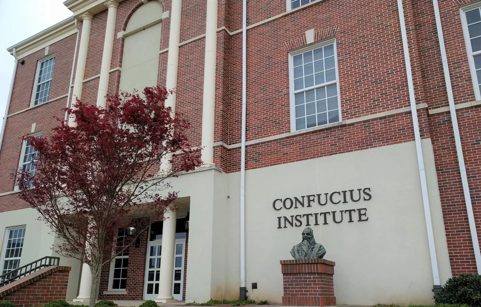 Кирпичное здание с белыми колоннами и надписью Confucius Institute на стене, перед ним бюст на постаменте и дерево с бордовой листвой