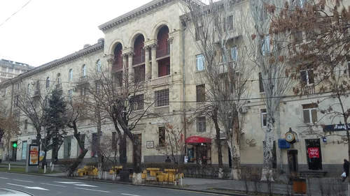 Pale tuff apartment block with arched balconies, cafés and a street clock among winter trees