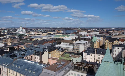 An elevated view over city-block rooftops with a white cathedral and the sea in the distance under a blue sky with clouds