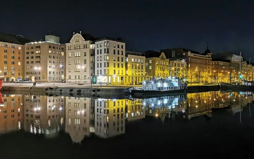 A row of lit buildings along a waterfront and a moored ship, their lights reflected in still water