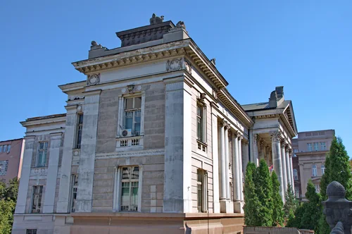 Pale colonnaded building with rooftop sculptures under a blue sky