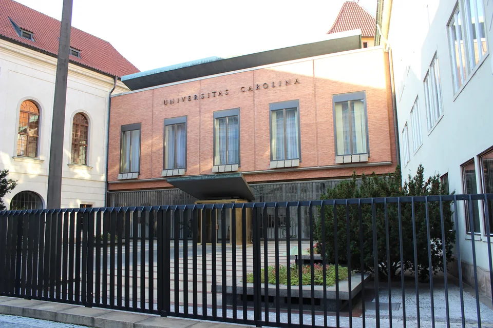 Кирпичный фасад с надписью «Universitas Carolina» за металлической оградой, рядом светлое здание с арочными окнами