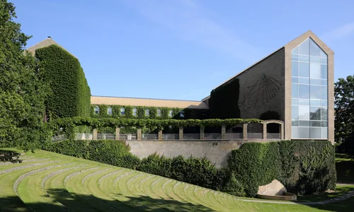 A yellow-brick building with ivy-covered walls and a tall glass window, with a grassy amphitheatre in front