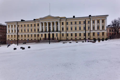 A yellow Neoclassical building with columns beyond a snow-covered square as snow falls