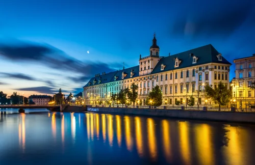A long Baroque building with a tower above a river, lit by street lamps whose lights reflect in the water under a twilight sky