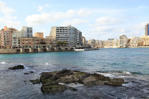 A rocky shore and turquoise sea, with rows of pale multi-storey buildings across the bay