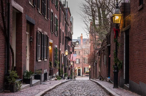 A narrow cobbled street between brick houses with dark shutters, street lamps and Christmas decorations