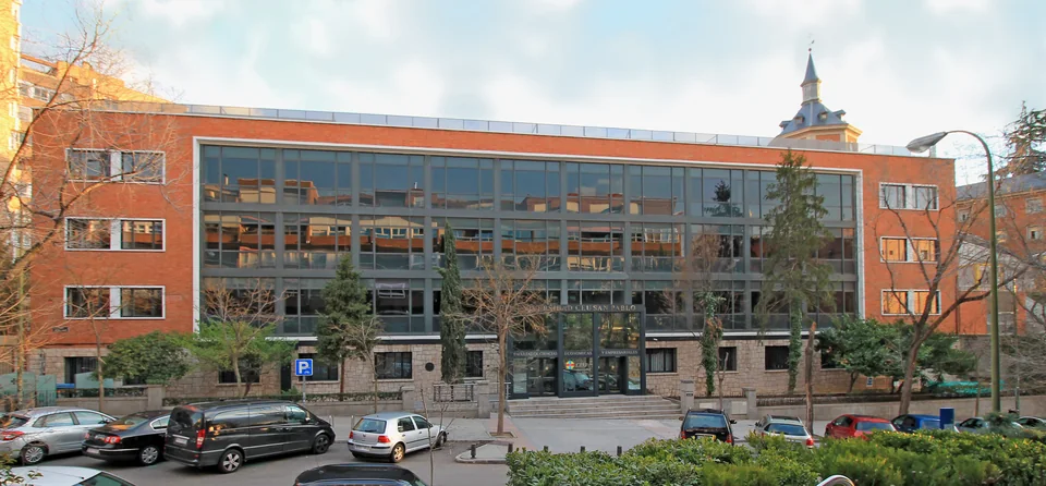 Стеклянный фасад между кирпичными крыльями с надписью Universidad CEU San Pablo над входом, перед ним парковка