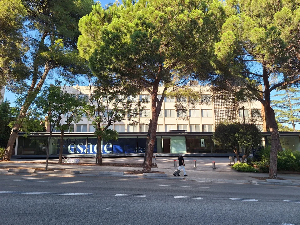 Остеклённый первый этаж с крупной надписью esade под многоэтажным корпусом, перед ним тротуар и деревья