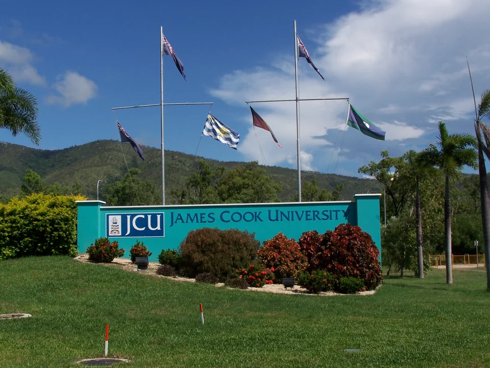 Бирюзовая стена с вывеской James Cook University среди цветущих кустов, над ней флагштоки с флагами, за ними горы