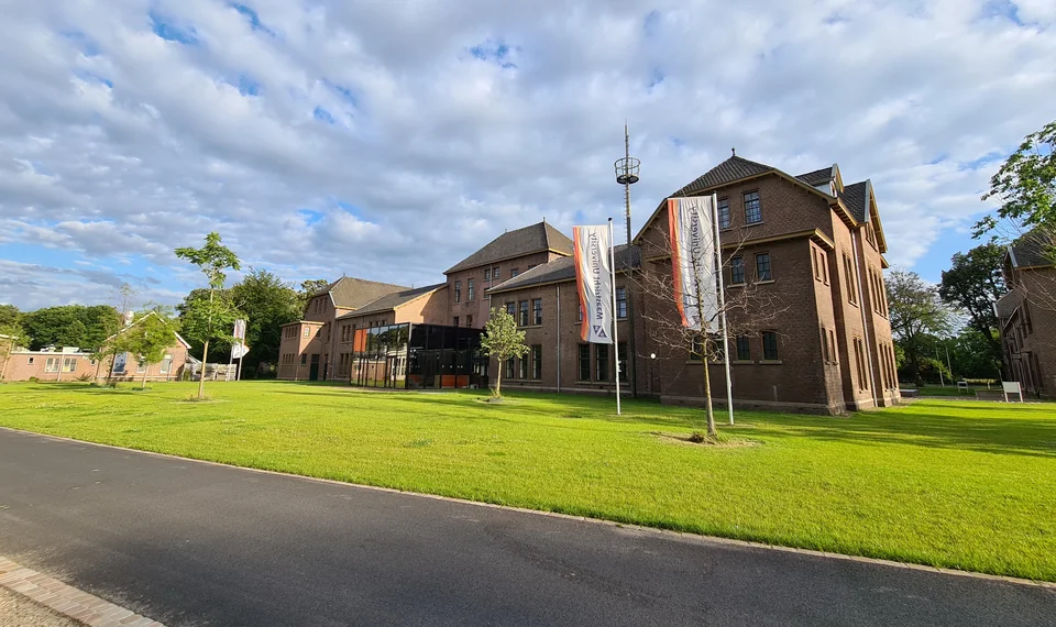 Кирпичные корпуса со стеклянной пристройкой и флагами Maastricht University за широким газоном