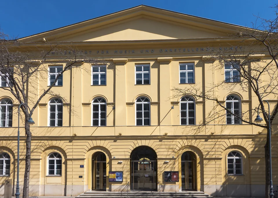Жёлтый классический фасад с фронтоном и надписью Universität für Musik und darstellende Kunst